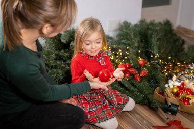 Young girl helping her mother decorating the Christmas tree, mother keeps some Christmas baubles in her hand. . High quality photo