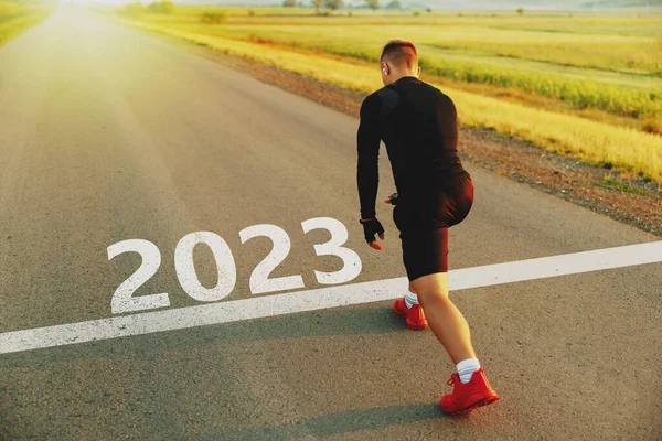 New year 2023 or start straight concept Word 2023 written on the asphalt road and athlete man runner stretching leg preparing for new year at sunset. Concept of challenge or career path or change.