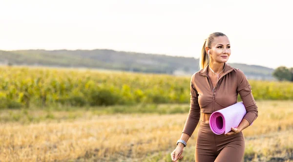 Beautiful woman walking through the field with yoga mat in hand with big smile looking for a place for recreation. High quality photo