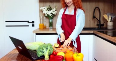 Kırmızı mutfak şapkalı güzel kız önünde bilgisayarla ayakta duran bir salata yapmak için sebze kesiyor. Yüksek kalite 4k görüntü