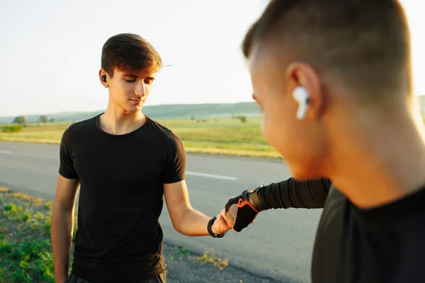 Muscular men clapping hands and preparing for workout an a field. Both listen to music on wireless headphones and wear a black t-shirt. High quality photo