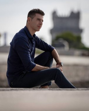 Portekiz 'de Lizbon nehri kıyısında tek başına dinlenen klas bir girişimcinin portresi. Şık bir iş adamı mavi şık bir gömlek ve resmi bir kıyafet giyiyor. Başarılı yaşam tarzı kavramı.