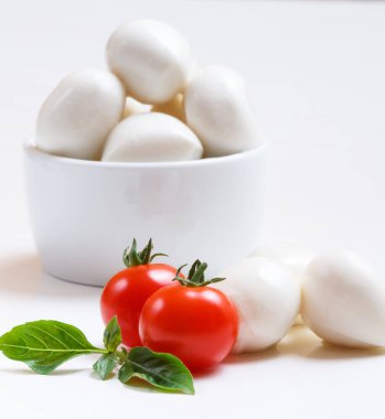 Beyaz kasede küçük mozzarella topları, kiraz domatesleri ve beyaz arka planda taze fesleğen.