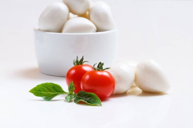 Beyaz kasede küçük mozzarella topları, kiraz domatesleri ve beyaz arka planda taze fesleğen.
