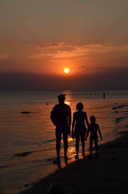 Sahilde gün batımını izleyen üç kişilik bir aile. Gün batımında üç kişi..