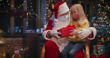 Koruyucu maske takan meraklı kız Noel Baba 'nın dizlerinin dibinde oturuyor ve alışılmadık hediyelerle hediye kutusunu karıştırıyor. Tatil kavramı