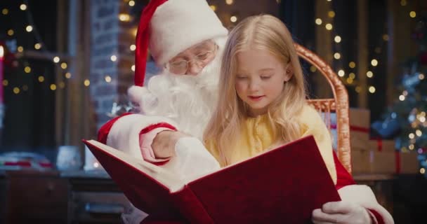 Szczęśliwa dziewczynka przytula Mikołaja i czyta książkę z zainteresowaniem. Szczęśliwe dziecko spędzające czas z Mikołajem. Koncepcja urlopu — Wideo stockowe