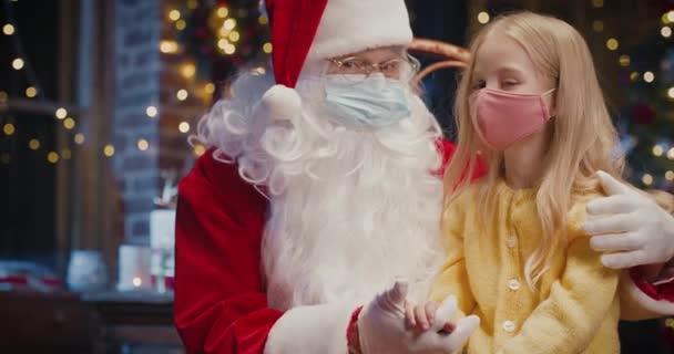 Portret van het kleine blonde meisje dat op de knieën van de Kerstman zit terwijl ze geheimen met hem deelt. Ze dragen beschermende maskers. Pandemisch concept — Stockvideo