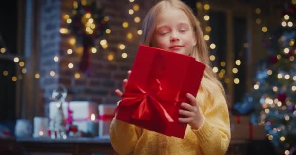 Kleine kaukasische Mädchen mit Weihnachtsmütze halten Geschenkbox und warten auf das Geschenk mit Weihnachtsbaum im Hintergrund — Stockvideo