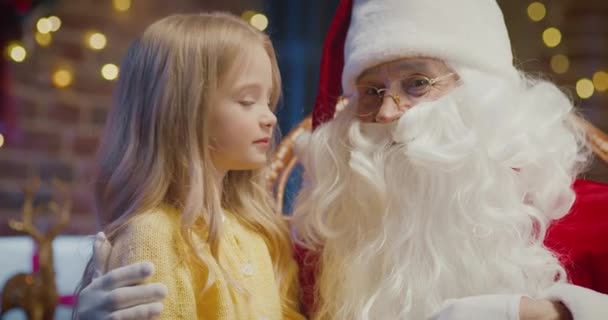 I will tell you but keep it in secret. Portrait view of the little blonde girl sitting at the Santa knees and looking at the camera while sharing secrets with him — Stock Video