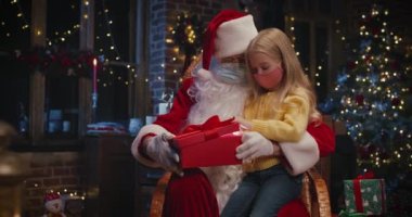 Koruyucu maske takan meraklı kız Noel Baba 'nın dizlerinin dibinde oturuyor ve alışılmadık hediyelerle hediye kutusunu karıştırıyor. Tatil kavramı