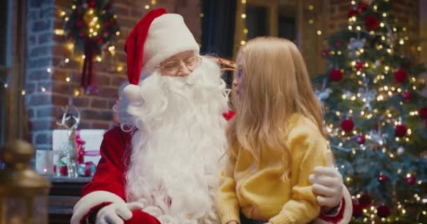 I will tell you but keep it in secret. Portrait view of the little blonde girl sitting at the Santa knees while sharing secrets with him — Stock Video