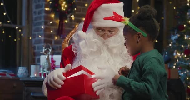 É para mim. Pequena menina multirracial de pé perto do Papai Noel e olhando para a caixa presente com interesse. Papai Noel olhando para ela com sorriso — Vídeo de Stock