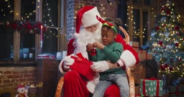 Noel Baba ve küçük çok kültürlü kız Noel Baba 'nın dizlerinin dibinde oturup Noel ağacının yanında kışın rahat oturma odasında sihirli bir Noel hediyesi bekliyorlar.