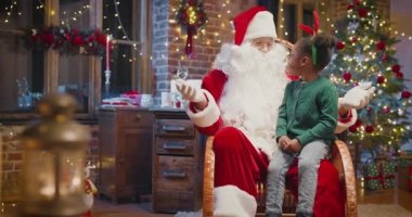 Noel Baba 'nın dizlerinin üzerine çöküp arka planda şöminesi olan mutlu, çok kültürlü bir kız.