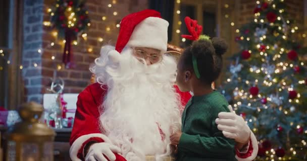 Happy little multiracial girl sitting on Santa Claus knees and telling something to him with fireplace in the background — Stock Video