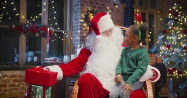 Küçük çok kültürlü kız Noel Baba 'nın dizlerinin önünde oturuyor ve hediyesini şu anki kutusundan faiziyle çıkarıyor. Noel Baba ona gülümseyerek bakıyor.