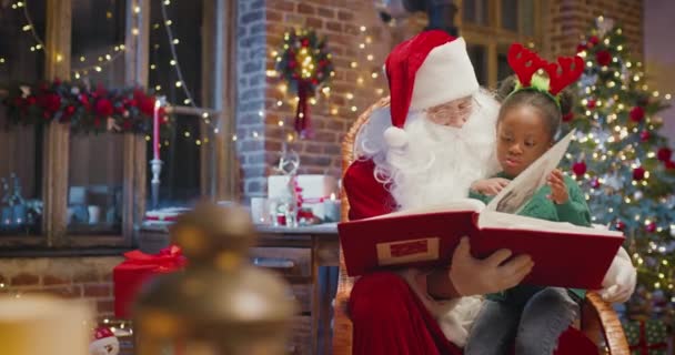 Счастливая многорасовая девочка обнимает Санту и с интересом читает книгу. Счастливого ребёнка, проводящего время с Санта Клаусом. Концепция праздников — стоковое видео