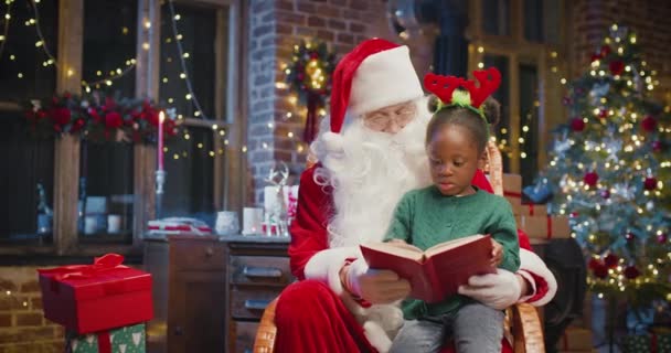 Menina criança multirracial feliz abraçando Papai Noel e livro de leitura com interesse. Criança feliz passando tempo com a Santa Cláusula. Conceito de feriados — Vídeo de Stock