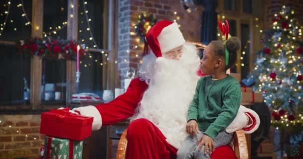 Küçük çok kültürlü kız Noel Baba 'nın dizlerinin önünde oturuyor ve hediyesini şu anki kutusundan faiziyle çıkarıyor. Noel Baba ona gülümseyerek bakıyor. — Stok video