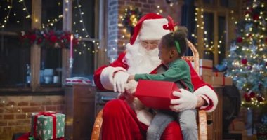 Küçük çok kültürlü kız Noel Baba 'nın dizlerinin önünde oturuyor ve hediyesini şu anki kutusundan faiziyle çıkarıyor. Noel Baba ona gülümseyerek bakıyor.