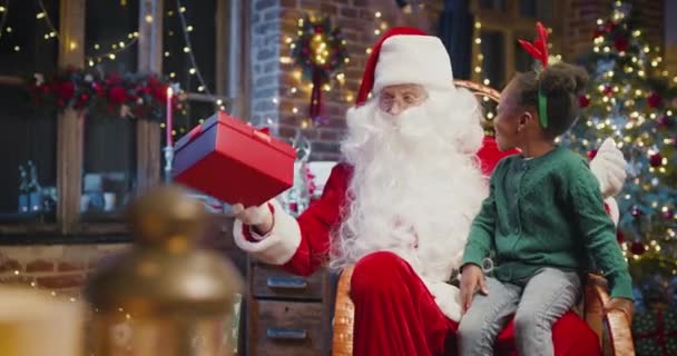 My Christmas present. Little multiracial girl sitting at the Santa knees and taking her gift from him with interest. Santa Claus looking at her with smile — Stock Video