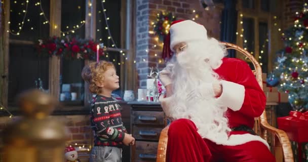 Mały biały chłopiec stojący obok Mikołaja i mówiący mu coś z przyjemnościami. Futra i dekoracje noworoczne w tle — Wideo stockowe