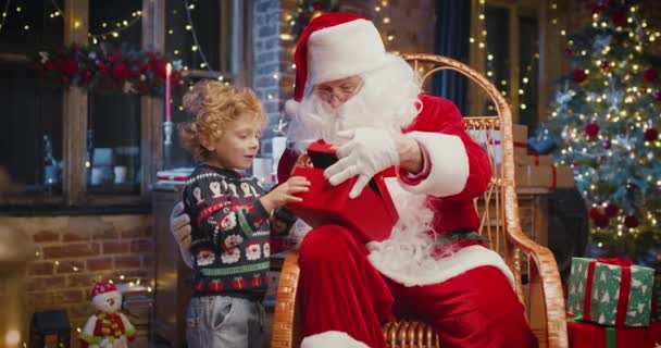 Mikulás és kis kaukázusi fiú áll a közelben, és várja a varázslatos karácsonyi ajándék közel a karácsonyfa hangulatos nappaliban télen — Stock videók