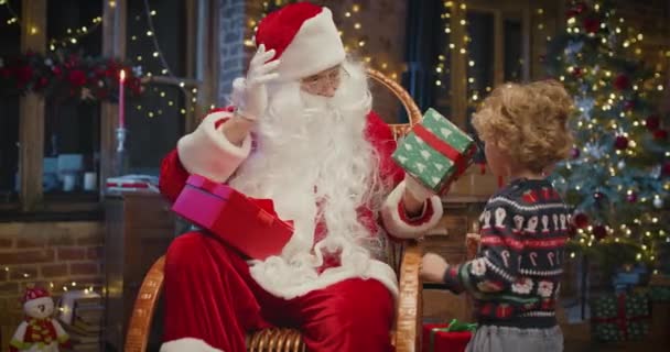 Санта Клаус з маленьким хлопчиком, який стоїть поруч і чекає чарівного різдвяного подарунка біля ялинки в затишній вітальні взимку. — стокове відео
