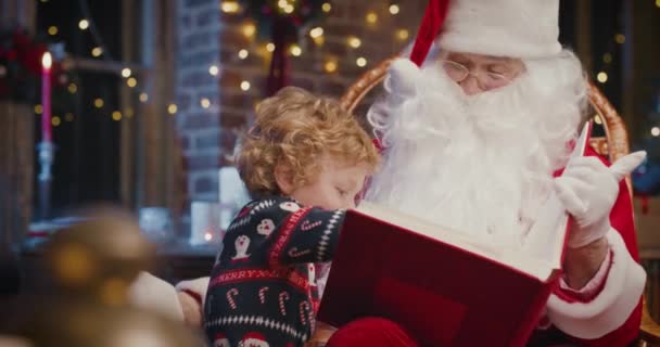 Glücklicher Junge, der den Weihnachtsmann umarmt und interessiert Bücher liest. Glückliches Kind, das Zeit mit dem Weihnachtsmann verbringt. Urlaubskonzept — Stockvideo