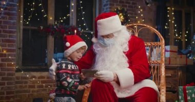 Küçük beyaz çocuk, Noel Baba 'yla akıllı telefonda çizgi film izlerken koruyucu maske takıyor ve yılbaşı süslemeleriyle evde vakit geçiriyor. Salgın konsepti