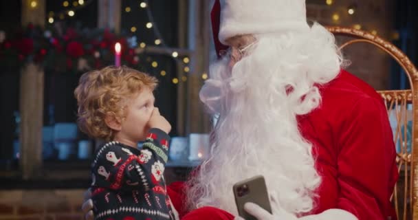 Petit garçon caucasien utilisant smartphone avec le Père Noël tout en passant du temps à la maison avec des décorations du Nouvel An. Concept de vacances — Video