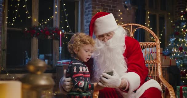 Med tomten. Liten kaukasiska pojke tittar på smartphone med jultomten samtidigt tillbringa tid hemma med nyår dekorationer — Stockvideo