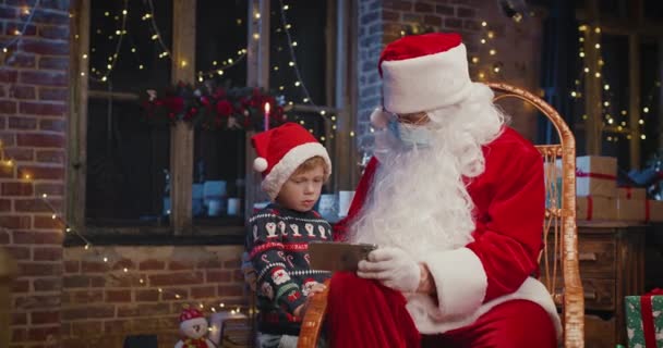 Pequeño chico caucásico usando teléfono inteligente con Santa Claus usando máscara protectora mientras pasa tiempo en casa con decoraciones de Año Nuevo. Concepto pandémico — Vídeos de Stock