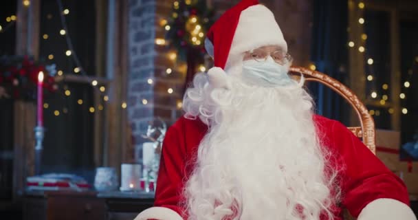 Hombre alegre Santa Claus usando máscara protectora mirando a la cámara mientras está sentado en casa. Primer plano del hombre mayor feliz disfrazado de Santa Claus. Concepto espíritu navideño — Vídeo de stock