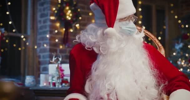 Święty Mikołaj w masce ochronnej siedzi na fotelu i pokazuje do symbolu serca aparatu siedząc w domu. Blisko szczęśliwego seniora w kostiumie Świętego Mikołaja. Koncepcja ducha Bożego Narodzenia — Wideo stockowe