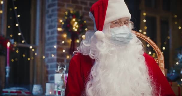 Gioioso uomo Babbo Natale indossa maschera protettiva guardando la fotocamera mentre seduto a casa. Primo piano di felice uomo anziano in costume da Babbo Natale. Concetto spirito natalizio — Video Stock