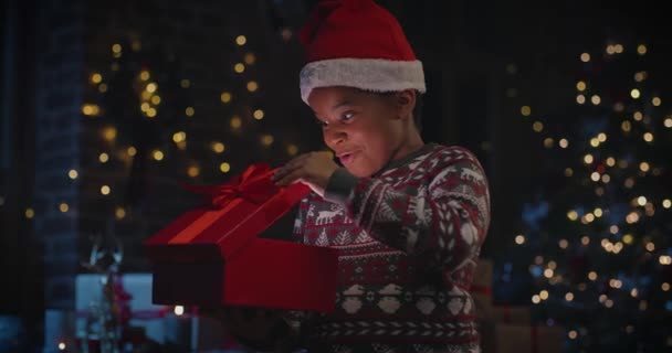 Kleine multiraciale kind jongen in Santa hoed openen geschenkdoos en zich afvragen van het geschenk met kerstboom op de achtergrond — Stockvideo