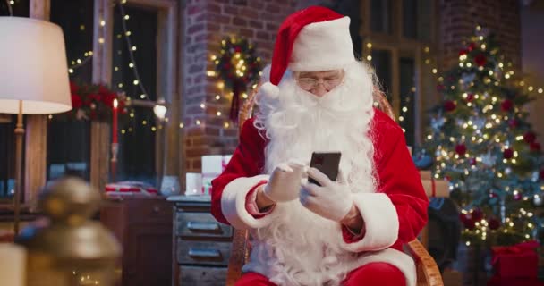 Santa Claus sentado en el sillón y el uso de teléfono inteligente mientras se sienta en casa con decoraciones de Año Nuevo. Concepto espíritu navideño — Vídeos de Stock