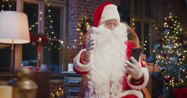 Der Weihnachtsmann sitzt am Sessel und bedient das Smartphone, während er zu Hause mit Neujahrsdekoration sitzt. Konzept zur Weihnachtsstimmung — Stockvideo