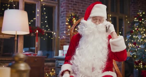 Закройте счастливого старшего человека в костюме Санта Клаузе, который болтает на смартфоне с кем-то, когда он сидит дома. Святкова концепція — стокове відео