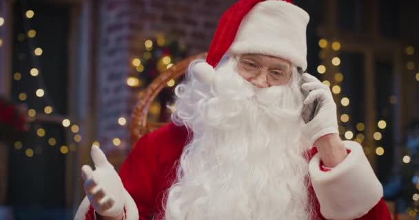 Närbild av glad senior man i Santa Clause kostym chatta på smartphone med någon när du sitter hemma. Begreppet semester — Stockvideo