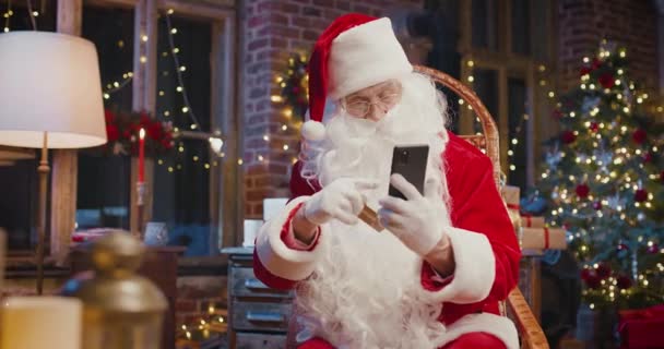 Iloinen valkohiuksinen joulupukki istuu nojatuolissa ja shoppailee luottokortilla älypuhelimella, jossa on monia joululahjoja ja lahjapakkauksia. Holidays henki käsite — kuvapankkivideo
