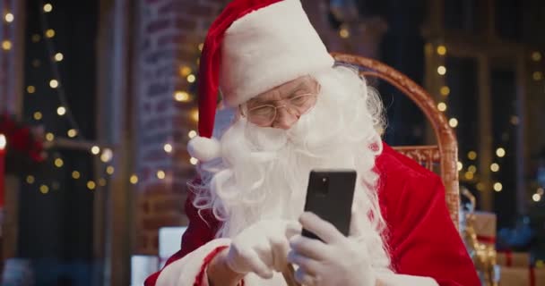 Glada vithåriga jultomten sitter vid fåtöljen och handlar med sitt kreditkort på smartphone med många julklappar och presentaskar runt. Begreppet semesteranda — Stockvideo