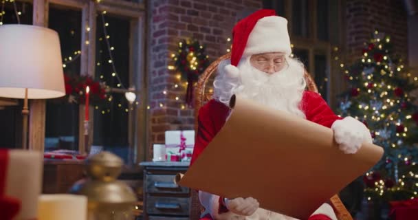 Babbo Natale barbuto seduto sulla poltrona ed esaminando carta con regali con decorazioni natalizie sullo sfondo. Vacanze spirito concetto — Video Stock