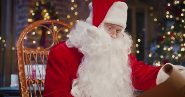 Babbo Natale barbuto seduto sulla poltrona ed esaminando carta con regali con decorazioni natalizie sullo sfondo. Vacanze spirito concetto — Video Stock