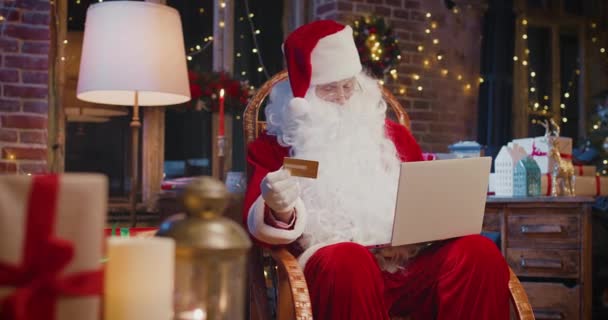 Wesoły siwowłosy Święty Mikołaj siedzi przy fotelu i robi zakupy z kartą kredytową na laptopie z wieloma prezentami świątecznymi i pudełkami z prezentami. Koncepcja świątecznego ducha — Wideo stockowe