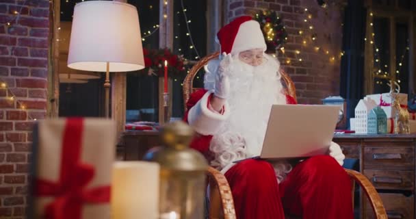 Noel Baba koltukta oturup dizüstü bilgisayar kullanırken yeni yıl süslemeleriyle evde oturuyor. Noel ruhu konsepti — Stok video