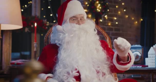 Санта Клаус сидит у кресла и смотрит в камеру с подарками с новогодними украшениями на заднем плане. Концепция праздничного духа — стоковое видео