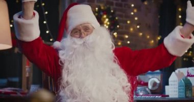 Noel Baba koltukta oturuyor ve evde otururken kameraya el sallıyor. Noel Baba kostümlü mutlu son sınıf öğrencisini kapatın. Noel ruhu konsepti